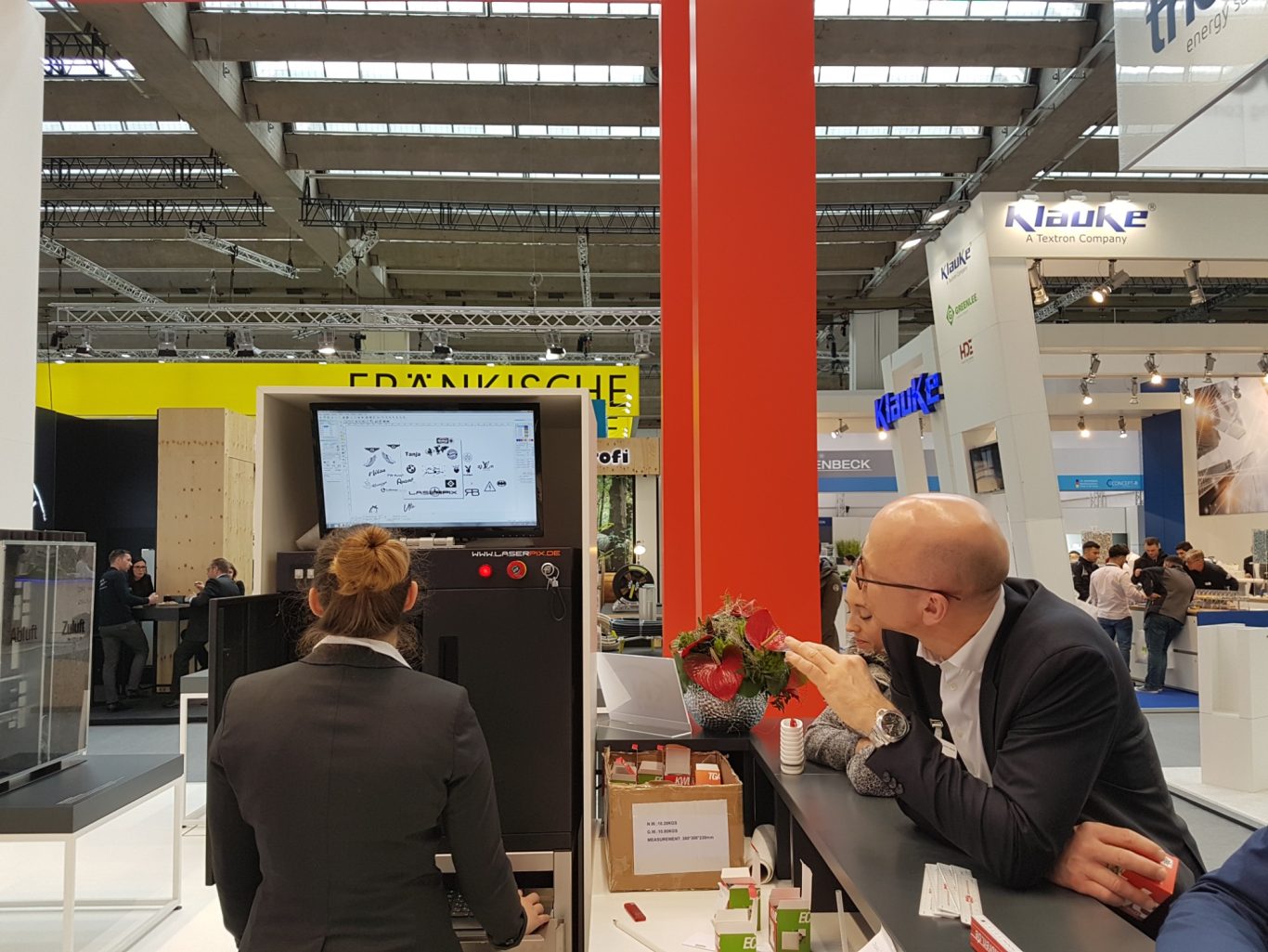 Messebesucher am Stand, eine Person sitzt vor einem Bildschirm, andere beobachtet.